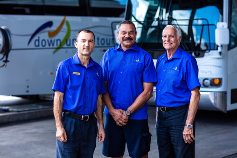 three tour guides in uniform standing smiling with tours buses in the background.